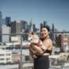 Cheerful Asian female in sportswear carrying cute puppy while walking together near city and looking at camera
