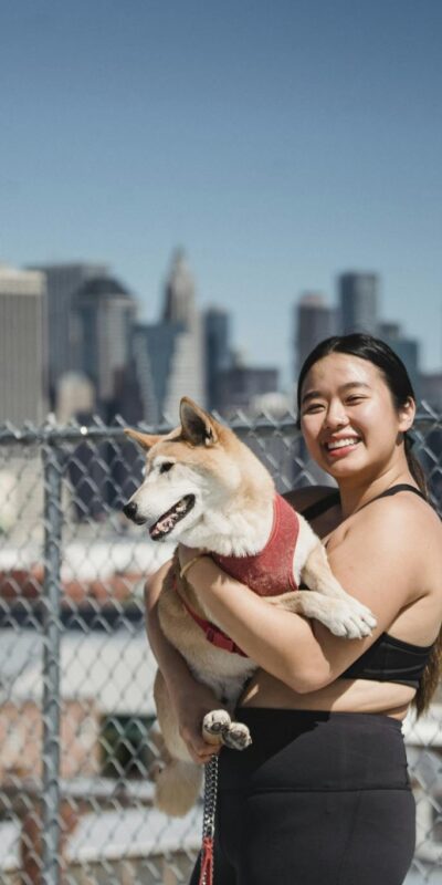 Cheerful Asian female in sportswear carrying cute puppy while walking together near city and looking at camera
